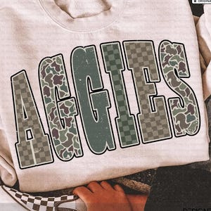 May include: A pale beige sweatshirt featuring the word "AGGIES" in large, block letters. The letters have a green outline, with the letters filled with a camouflage and checkered pattern. The text "DESIGNED BY DESIGNWINCO" is visible.