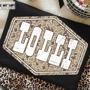 May include: Black t-shirt with a distressed, camouflage-patterned graphic that reads "LOLII" in white block letters. The shirt is folded, and a leopard print fabric is visible at the bottom. The design includes the text "designwinco DO NOT COPY ORIGINAL DESIGN".
