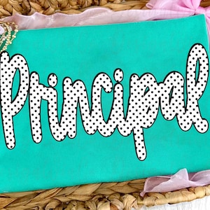 May include: A turquoise t-shirt with the word "Principal" in large, white, polka-dotted letters. The shirt is displayed on a woven basket with pink and gold accents.
