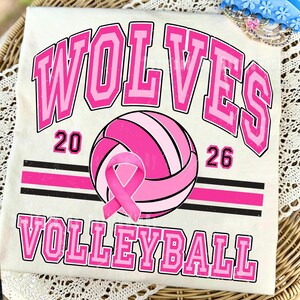 May include: A cream-colored t-shirt featuring a pink and black graphic design. The text reads WOLVES 20 26 VOLLEYBALL. A pink volleyball is centered with a pink awareness ribbon overlay. The design includes horizontal stripes and a distressed texture.