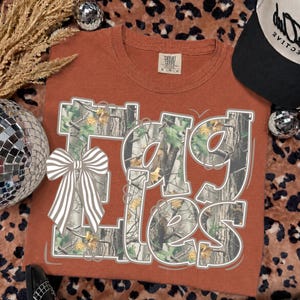 May include: A rust-colored short-sleeve t-shirt featuring the text "EAGLES" in a large, blocky font filled with a camouflage pattern of tree bark and green leaves. A white and gray striped bow graphic is positioned over the letter E.