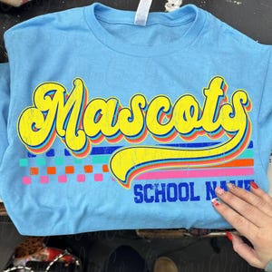 May include: A light blue t-shirt with the word "Mascots" in yellow, outlined in blue and red. Below the word is a horizontal stripe design with pink, blue, and green squares. The words "SCHOOL NAME" are below the stripe.