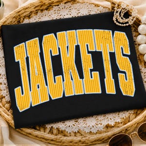 May include: A black t-shirt featuring the word JACKETS in a large, arched, yellow font with a distressed, vertical-striped texture and a thin white outline. The lettering is bold and follows a collegiate-style design across the front.