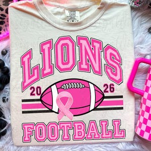 May include: A cream-colored t-shirt featuring the text LIONS in large pink block letters above a pink football graphic with a pink awareness ribbon. The numbers 20 and 26 flank the football, and the word FOOTBALL appears in pink below the graphic.
