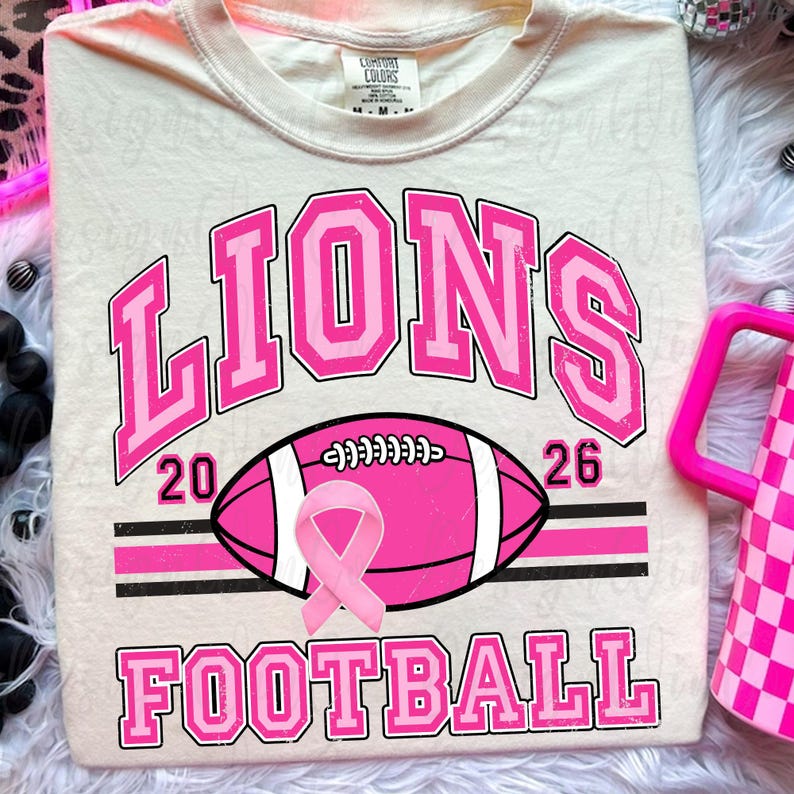 May include: A cream-colored t-shirt featuring the text LIONS in large pink block letters above a pink football graphic with a pink awareness ribbon. The numbers 20 and 26 flank the football, and the word FOOTBALL appears in pink below the graphic.