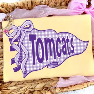Könnte beinhalten: Ein hellgelbes T-Shirt mit einer lila-weiß karierten Wimpelgrafik und dem Text "Tomcats" in fetten lila Buchstaben. Das Design enthält eine große lila Schleife, die an der Seite des Wimpels befestigt ist und einen mehrschichtigen Look erzeugt.