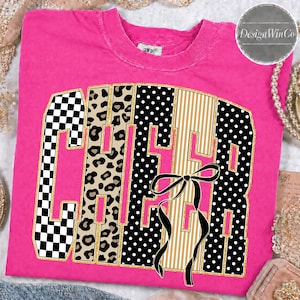 May include: A hot pink short-sleeved t-shirt featuring the word CHEER in large block letters. Each letter has a different pattern: black and white checkerboard, leopard print, black with white polka dots, and tan with white vertical stripes.