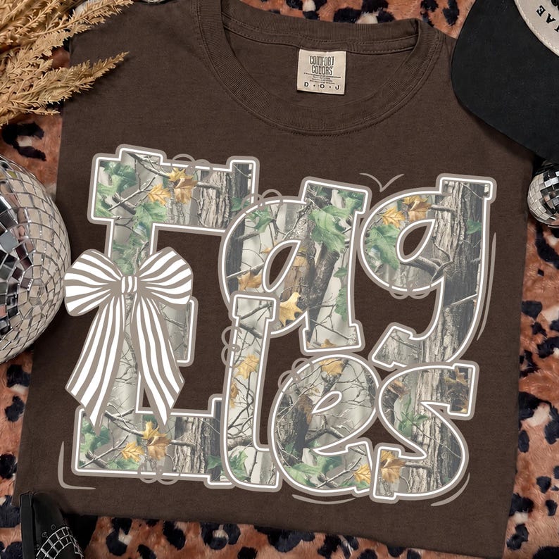 May include: A dark brown t-shirt featuring the text "EAGLES" in large, blocky letters filled with a camouflage pattern of tree bark and green and yellow leaves. A large, light-colored bow with white stripes is positioned over the letter E.