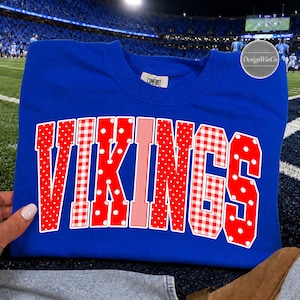 Peut inclure: Un t-shirt à col rond bleu roi avec le mot VIKINGS en grandes lettres style bloc. Les lettres sont rouges avec un contour blanc et présentent des motifs alternés de pois blancs, de carreaux vichy rouges et blancs, et de rayures verticales.
