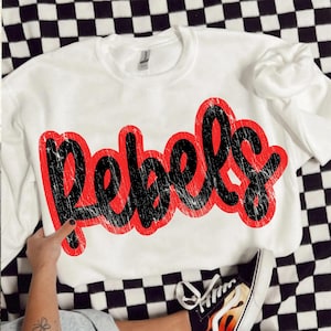 May include: White sweatshirt with the word "Rebels" in large, distressed black lettering with a red outline. The sweatshirt is on a black and white checkered surface.