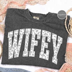 May include: A charcoal gray short-sleeve t-shirt with a crew neckline. The front features the word WIFEY in large, block-style varsity lettering. The letters have a white and gray leopard print pattern with a thin white outline.
