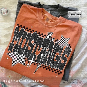 May include: An orange t-shirt with the word "MUSTANGS" in a bold, stylized font with a leopard print pattern. The design includes checkered stars and lightning bolts. A gray t-shirt is visible in the background.