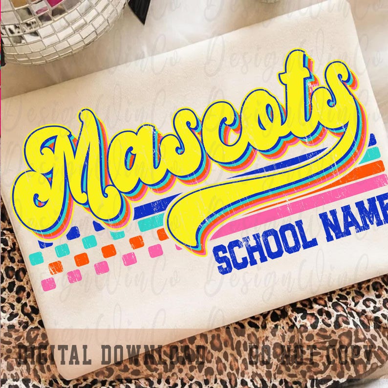 May include: Cream-colored t-shirt with the word "Mascots" in large, yellow, retro-style lettering with blue and red outlines. Below the word are horizontal stripes in pink, blue, and orange. The words "SCHOOL NAME" are below the stripes in blue.