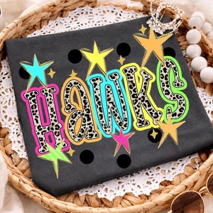 May include: A dark gray t-shirt featuring the word HAWKS in a leopard print pattern with colorful outlines. The design is accented with various stars in yellow, orange, pink, and turquoise, along with several solid black circular dots.