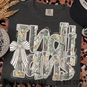 Könnte beinhalten: Ein anthrazitfarbenes T-Shirt mit dem Schriftzug "Indi Hans" in großen, fetten Buchstaben, die mit einem Wald-Tarnmuster aus Bäumen und Blättern gefüllt sind. Eine weiß-grau gestreifte Schleife ist an der linken Seite des Buchstabens I befestigt.