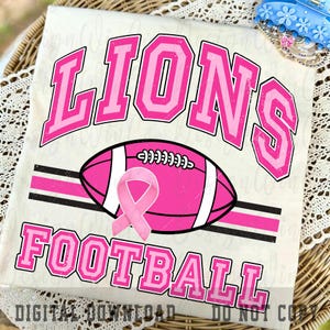 May include: A white t-shirt featuring the word LIONS in large pink varsity letters above a pink football graphic with a pink awareness ribbon. The word FOOTBALL is printed in pink varsity letters below the football, with black horizontal stripes.