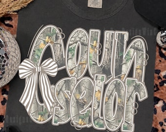 May include: A charcoal gray t-shirt featuring the text "COUNSELOR" in large, bold letters filled with a forest camouflage pattern of trees and leaves. A white and gray striped bow is attached to the left side of the letter C. The shirt has a Comfort Colors brand label.