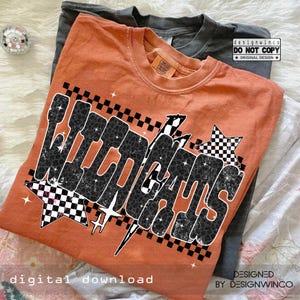 Pode incluir: Uma camiseta laranja com a palavra "WILDCATS" em preto, com uma fonte de estampa de leopardo, acentuada com estrelas quadriculadas e um raio. A camiseta est&aacute; dobrada sobre uma camiseta cinza. As palavras "digital download" est&atilde;o na parte inferior.