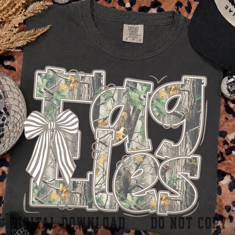 May include: A charcoal gray t-shirt featuring the word EAGLES in large, bold, white-outlined letters filled with a camouflage pattern of tree bark and green leaves. A white and gray striped bow is positioned over the letter E on the left side.