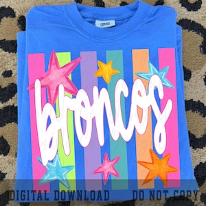 May include: A blue t-shirt with the word "Broncos" in white script, set against a backdrop of vertical stripes in various colors. The design includes colorful star accents, creating a vibrant and playful look. The shirt is folded.