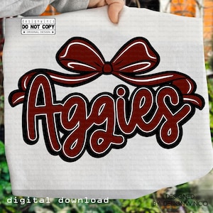 May include: A maroon and black graphic with the word "Aggies" in a cursive font, outlined in black, and a maroon bow at the top. The words "designwinco" and "digital download" are also visible.
