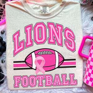 May include: A cream-colored t-shirt featuring the word LIONS in pink varsity-style lettering above a pink football graphic with a pink awareness ribbon. The word FOOTBALL is printed in pink below the football, all set against a light background.