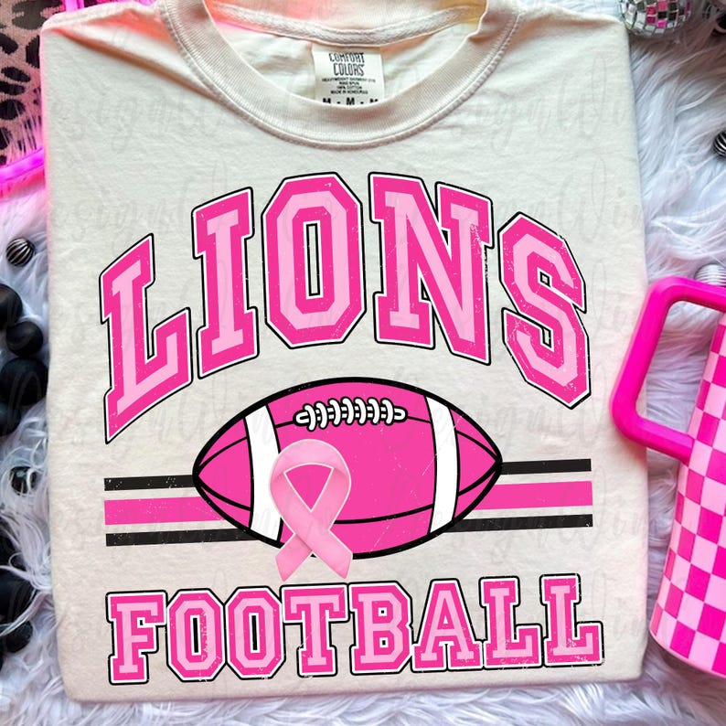 May include: A cream-colored t-shirt featuring the word LIONS in pink varsity-style lettering above a pink football graphic with a pink awareness ribbon. The word FOOTBALL is printed in pink below the football, all set against a light background.