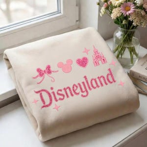 May include: A folded beige blanket with pink embroidered Disneyland graphics. The design includes a bow, Mickey Mouse silhouette, heart, castle, stars, and the word "Disneyland" in a glittery font.