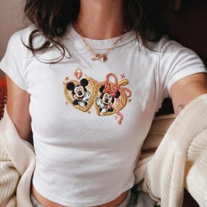 May include: White t-shirt with embroidered Mickey and Minnie Mouse design. The design features Mickey and Minnie in heart-shaped frames with gold and pink accents. A gold necklace with a heart pendant is also visible.