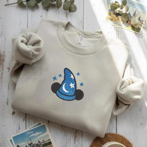 Op de afbeelding: Een lichtbeige sweatshirt met een blauw tovenaars hoed ontwerp, met een maan, sterren en Mickey Mouse oren. Het ontwerp is geborduurd met glitter. Het sweatshirt is op een houten oppervlak gevouwen.