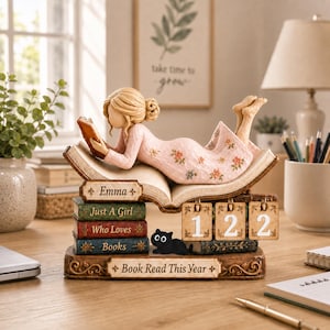 May include: A decorative figurine featuring a person with blonde hair in a bun wearing a light pink floral dress, lying on an open book. The base includes stacked books labeled Emma, Just A Girl, Who Loves Books, and Book Read This Year, with a black cat and a calendar displaying 122.