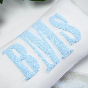 May include: White rectangular pillow with the embroidered letters "BMS" in light blue. The letters have a textured, raised appearance. The pillow is on a white surface.