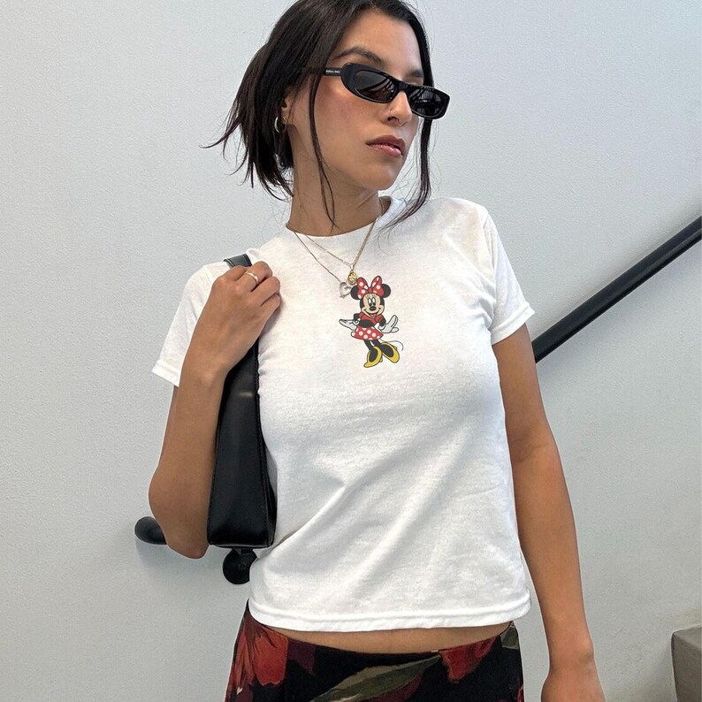 Puede incluir: Camiseta blanca con un estampado de Minnie Mouse. El personaje de dibujos animados lleva un vestido de lunares rojos y blancos, zapatos amarillos y un lazo rojo. La camiseta se combina con un bolso negro y gafas de sol.
