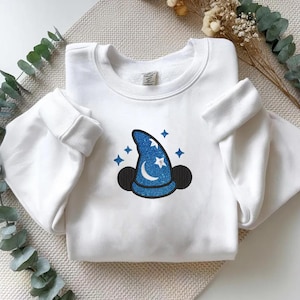 Op de afbeelding: Witte sweatshirt met een blauw glitter tovenaars hoed ontwerp, met een maan, sterren en zwarte muizenoren. Het ontwerp staat op de voorkant. De mouwen hebben manchetten.