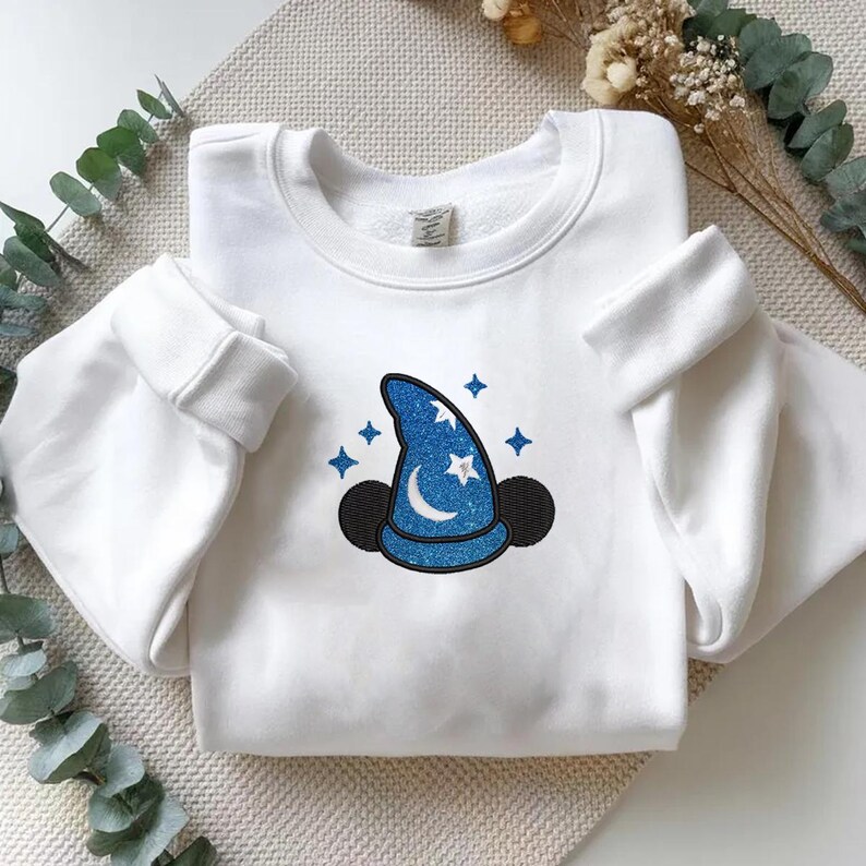 Op de afbeelding: Witte sweatshirt met een blauw glitter tovenaars hoed ontwerp, met een maan, sterren en zwarte muizenoren. Het ontwerp staat op de voorkant. De mouwen hebben manchetten.