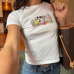 May include: White t-shirt with a colourful "Disneyworld" graphic featuring Mickey Mouse, Minnie Mouse, Donald Duck, Daisy Duck, Goofy, and Pluto. The shirt is paired with blue jeans. A brown handbag is held in the right hand.