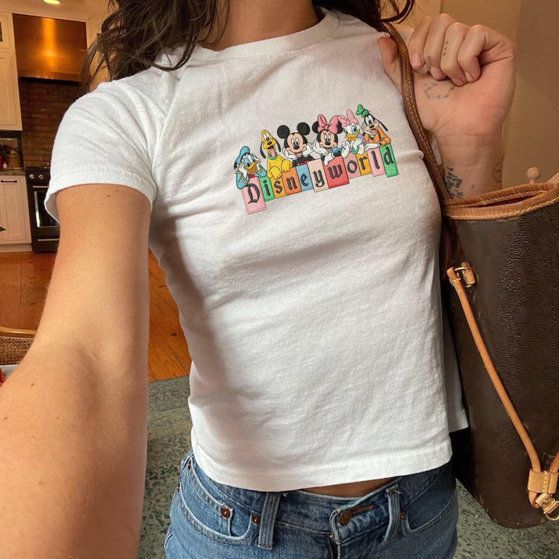 May include: White t-shirt with a colourful "Disneyworld" graphic featuring Mickey Mouse, Minnie Mouse, Donald Duck, Daisy Duck, Goofy, and Pluto. The shirt is paired with blue jeans. A brown handbag is held in the right hand.