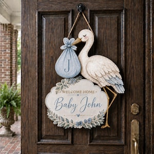 May include: A decorative wooden door hanger shaped like a stork carrying a blue bundle with the letter J. Below the stork is an oval sign with floral accents that reads Welcome Home Baby John in blue script and serif lettering.
