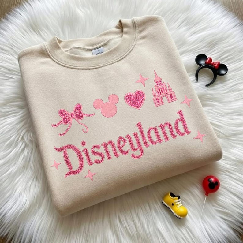May include: A beige sweatshirt with the word "Disneyland" in pink glitter lettering. The design includes a bow, Mickey Mouse head, heart, castle, and stars. Accessories include a Minnie Mouse ear headband, a yellow shoe, and a red balloon.