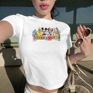 May include: White crop top with a "Disneyworld" graphic featuring cartoon characters. The design includes Mickey Mouse, Minnie Mouse, Donald Duck, and Goofy. The shirt is short-sleeved and made of a soft material.