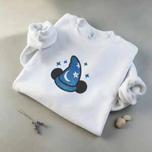 Op de afbeelding: Witte sweatshirt met een blauw glitter tovenaars hoed ontwerp, een maan, sterren en zwarte muizenoren. Het ontwerp is geborduurd op de voorkant. De mouwen zijn opgerold.