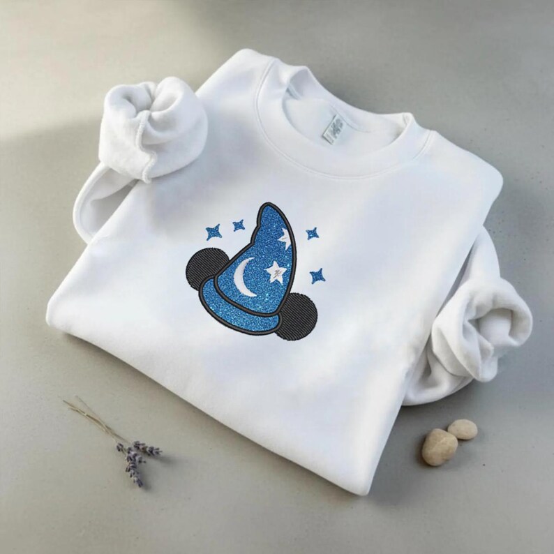 Op de afbeelding: Witte sweatshirt met een blauw glitter tovenaars hoed ontwerp, een maan, sterren en zwarte muizenoren. Het ontwerp is geborduurd op de voorkant. De mouwen zijn opgerold.