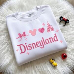 May include: White sweatshirt with pink Disneyland graphic. The design includes a bow, Mickey Mouse head, heart, castle, and stars. Accessories include a Minnie Mouse ear headband, a yellow shoe, and a red balloon.