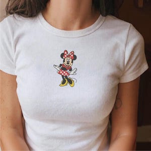 Puede incluir: Camiseta blanca con un estampado de Minnie Mouse. El personaje de dibujos animados lleva un vestido de lunares rojos y blancos, un lazo a juego y zapatos amarillos. La camiseta tiene cuello redondo y mangas cortas.
