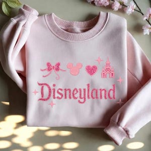 May include: A light pink sweatshirt with the word "Disneyland" in glittery pink letters. The design includes a bow, Mickey Mouse ears, a heart, a castle, and stars, all in pink.