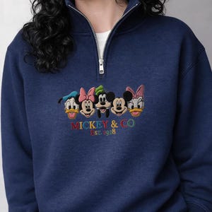 以下が含まれることがあります： ネイビーブルーの襟付きスウェットシャツ。前面には、ドナルドダック、ミニーマウス、ミッキーマウス、デイジーダックのディズニーキャラクターの刺繍デザインが施されています。「MICKEY & CO EST. 1928」の文字が下に刺繍されています。