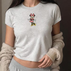 Puede incluir: Una camiseta gris claro con un dise&ntilde;o de Minnie Mouse. El dise&ntilde;o muestra a Minnie Mouse con un vestido rojo con lunares blancos, un lazo rojo y zapatos amarillos. La camiseta tiene mangas cortas y cuello redondo.
