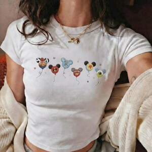 May include: White t-shirt with a crew neck featuring a colorful graphic of cartoon character-themed balloons. The balloons have various designs, including a cowboy hat, a superhero mask, and checkered patterns. The shirt is short-sleeved.