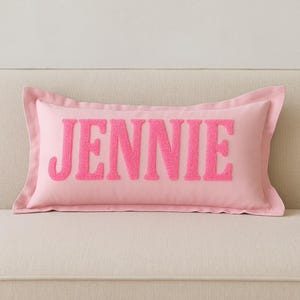 Op de afbeelding: Een roze rechthoekig kussen met de naam "JENNIE" geborduurd in een getextureerde, felroze lettertype. Het kussen heeft een zachte, lichtroze rand en ligt op een lichtgekleurde bank.