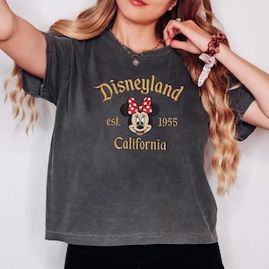 May include: Dark gray t-shirt with the Disneyland logo embroidered in gold thread. The design includes the text "Disneyland est. 1955 California" and a Minnie Mouse graphic with a red bow. The shirt has short sleeves.